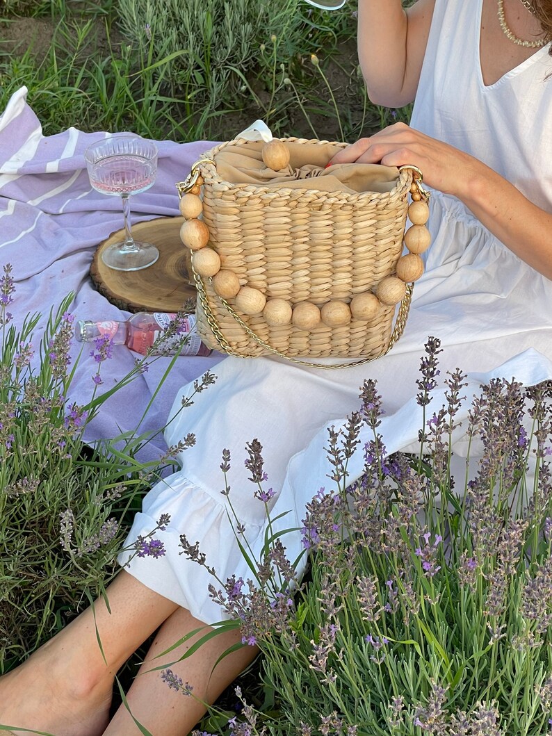 May include: A woven straw bag with a wooden bead handle. The bag is light brown and has a natural, rustic look. The bag is sitting on a purple blanket with lavender flowers in the background.