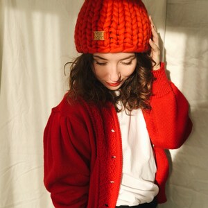 May include: A vibrant red, chunky knit beanie hat with a matching pom-pom. The hat features a small, rectangular label that reads "Your Yarn". The beanie is worn with a red cardigan. The cardigan has a button-up front and a textured knit design.