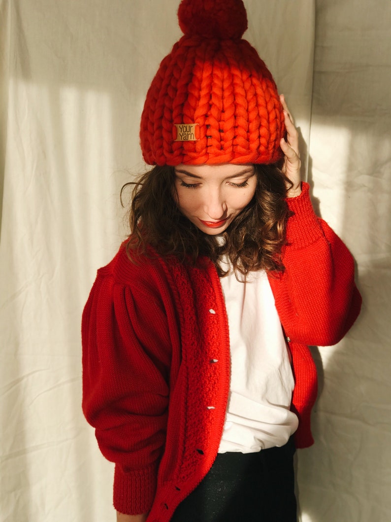 May include: A vibrant red, chunky knit beanie hat with a matching pom-pom. The hat features a small, rectangular label that reads "Your Yarn". The beanie is worn with a red cardigan. The cardigan has a button-up front and a textured knit design.