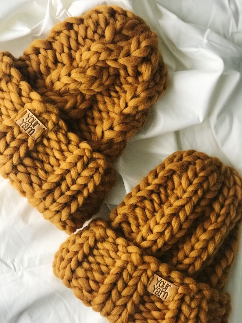 May include: Two mustard yellow knitted hats with a cable knit pattern. Each hat has a brown leather tag with the text "Your Yarn" on it.
