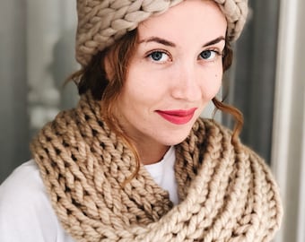 May include: A woman wearing a beige knitted headband and a matching scarf. The headband is a thick, chunky knit with a twisted design. The scarf is also a thick, chunky knit and is wrapped around the woman's neck.
