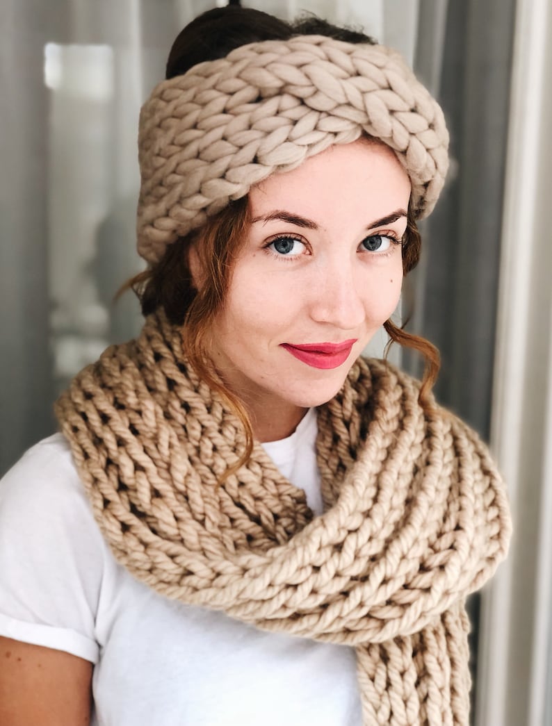 May include: A woman wearing a beige knitted headband and a matching scarf. The headband is a thick, chunky knit with a twisted design. The scarf is also a thick, chunky knit and is wrapped around the woman's neck.