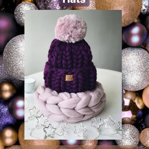 May include: A purple knitted beanie with a fluffy pink pom pom and a brown leather tag that says "Your Yarn". The beanie is sitting on a thick, light pink knitted circle. The background is a collection of sparkly purple, gold, and silver ornaments.
