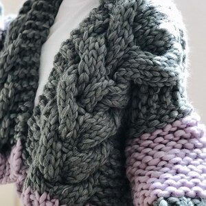May include: A chunky knit cardigan with a cable knit pattern in grey and purple. The cardigan has a V-neck and long sleeves.