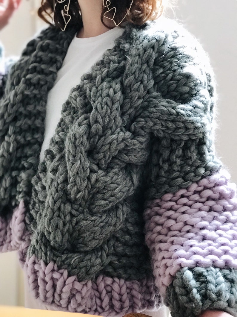 May include: A chunky knit cardigan with a cable knit pattern in grey and purple. The cardigan has a V-neck and long sleeves.