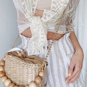May include: A straw bag with wooden beads around the top and a gold chain strap. The bag is worn over the shoulder of a person wearing a white lace top and striped shorts.
