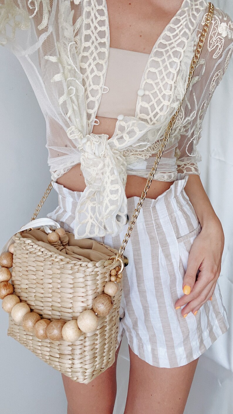 May include: A straw bag with wooden beads around the top and a gold chain strap. The bag is worn over the shoulder of a person wearing a white lace top and striped shorts.
