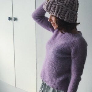 May include: A woman wearing a lavender fuzzy sweater and a silver skirt. She is wearing a chunky knit lavender beanie with a pom pom on top.