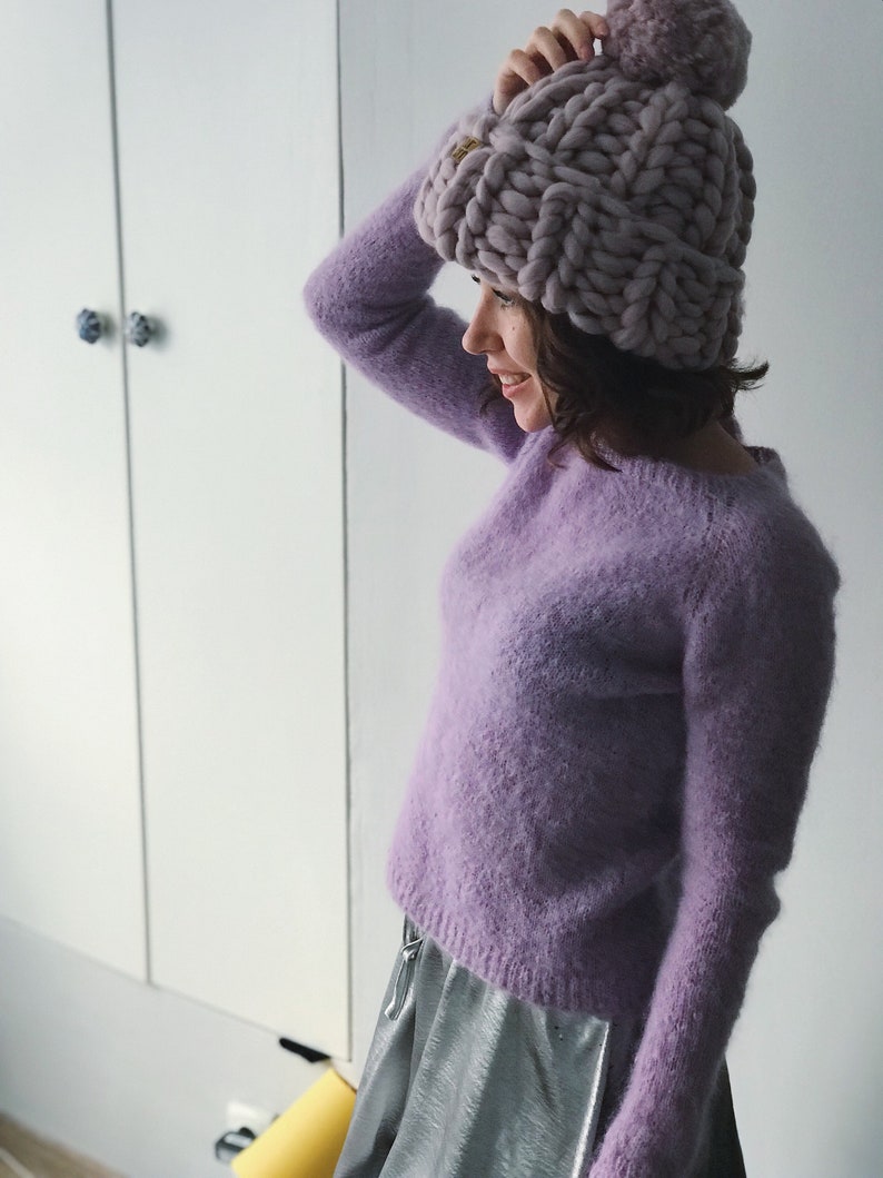 May include: A woman wearing a lavender fuzzy sweater and a silver skirt. She is wearing a chunky knit lavender beanie with a pom pom on top.