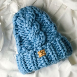 May include: A blue knitted beanie with a cable knit pattern and a brown leather tag that says "Your Yarn".