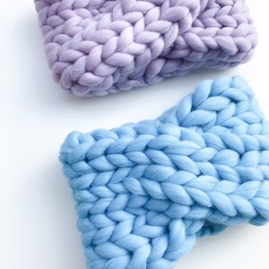 May include: Two chunky knit headbands, one in a light purple and one in a light blue. The headbands are made with thick yarn and have a simple, classic design.