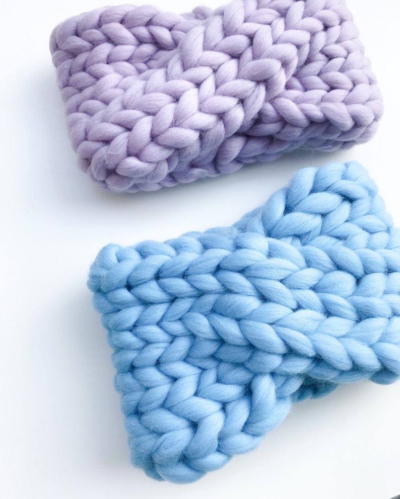 May include: Two chunky knit headbands, one in a light purple and one in a light blue. The headbands are made with thick yarn and have a simple, classic design.