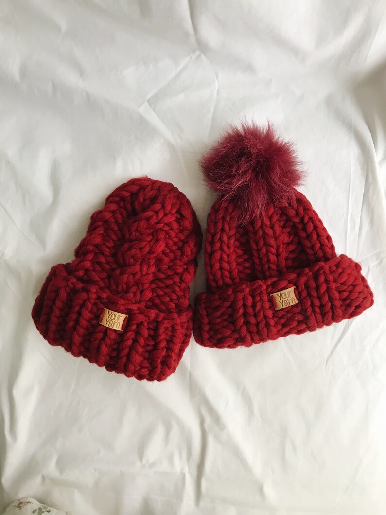 May include: Two red knitted beanies with pom poms. One beanie has a "Your Yarn" label.