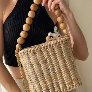May include: A straw tote bag with a wooden bead handle and a gold chain strap. The bag is woven with a light brown straw and has a rectangular shape.
