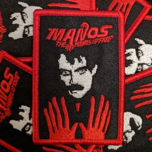 May include: A black and red embroidered patch with the text "MANOS THE HANDS OF FATE" and a white silhouette of a person's face with a mustache. The person's hands are shown in red below the face.
