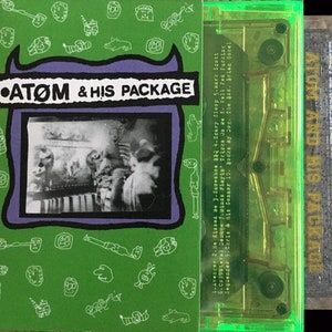 May include: A green cassette tape with a purple and black label that says "ATOM & HIS PACKAGE". The cassette tape is in a clear plastic case with a green border. The label has a black and white photo of a person in a room.