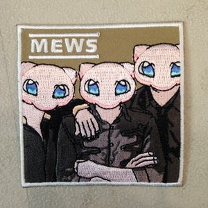 May include: Embroidered patch featuring three cartoon figures with pink cat-like faces and large blue eyes, dressed in dark clothing. The word "MEWS" is embroidered in white block letters above the figures on a tan background. The patch has a white border.