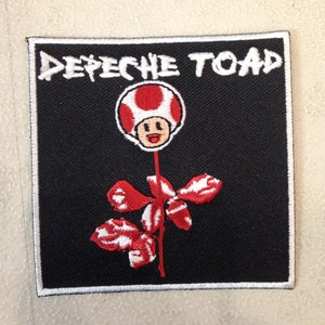 May include: Black embroidered patch with the text "Depeche Toad" in white and a red and white mushroom with a face on a stem with red leaves.