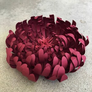 K&ouml;nnte beinhalten: Eine gro&szlig;e, handgefertigte Papierblume in Burgundert&ouml;nen. Die Blume hat geschichtete Bl&uuml;tenbl&auml;tter, die ein volles, strukturiertes Aussehen erzeugen. Die Mitte ist ein hellerer Rotton. Ideal f&uuml;r Heimdekoration oder besondere Anl&auml;sse.