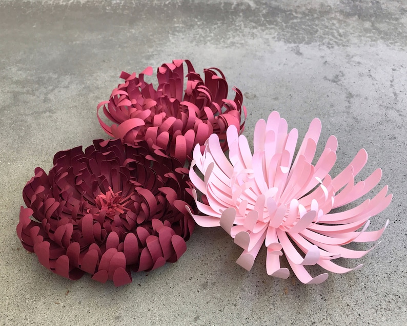 K&ouml;nnte beinhalten: Drei Papierblumen in Rot- und Rosat&ouml;nen. Die Blumen sind aufwendig geschnitten und geschichtet, mit einem detaillierten, strukturierten Aussehen. Die Blumen sind auf einer grauen Oberfl&auml;che angeordnet und bilden eine ansprechende Darstellung.
