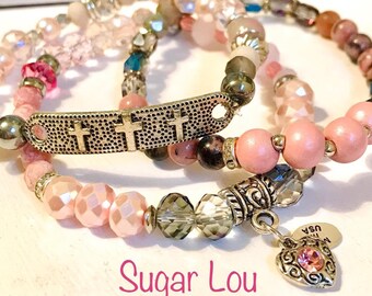 Könnte beinhalten: Mehrere Armbänder mit Perlen in Rosa, Grau und Transparent. Ein Armband hat einen silbernen Balken mit drei Kreuzen. Ein anderes hat einen Herzanhänger mit einem rosa Edelstein. Der Text "Sugar Lou" steht unten.