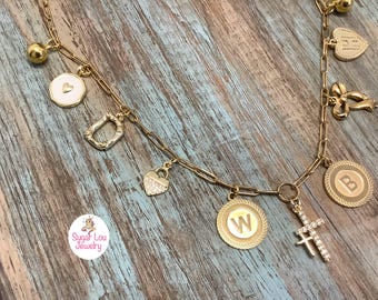 Puede incluir: Collar de dijes dorado con varios colgantes, incluyendo un corazón, una cruz, un lazo y medallones con las letras "W" y "B". El collar tiene una cadena delicada y se muestra sobre una superficie de madera rústica.