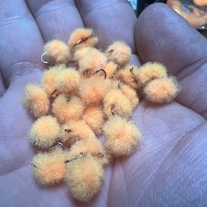 May include: A collection of peach-colored fly fishing lures with gold hooks. The lures are made of fluffy material and are arranged in a hand.