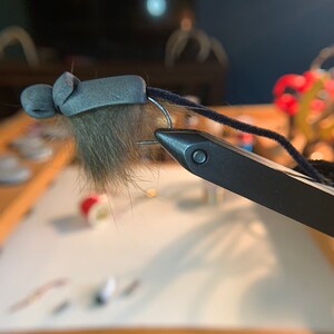 May include: A close-up of a fly tying vise holding a gray and brown fly with a black thread wrapped around the hook.