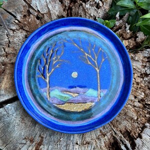 May include: A round ceramic plate with a blue rim and a central landscape design. The design features a blue sky with a full moon, two stylized trees, and rolling hills in shades of purple, green, and tan.