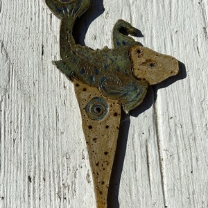 May include: A ceramic fish-shaped art piece with a speckled, tan body and green and blue accents. The fish has a detailed tail and a pointed base. The artwork is displayed against a white, textured background.