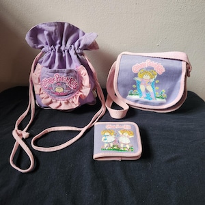 May include: A collection of vintage Cabbage Patch Kids accessories, including a drawstring bag, a small purse, and a wallet. The items are primarily lavender and pink, with the Cabbage Patch Kids logo and character illustrations.