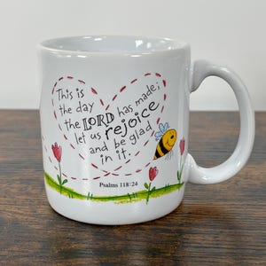 Puede incluir: Una taza de cerámica blanca con un contorno de corazón dibujado a mano en líneas discontinuas rojas que contiene el texto: This is the day the LORD has made; let us rejoice and be glad in it. Psalms 118:24. El diseño incluye una pequeña abeja amarilla y negra y flores rojas.