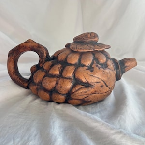 May include: A brown ceramic teapot with a textured, organic design. The teapot has a lid, a curved handle, and a spout. The body of the teapot has a unique, textured surface, and the overall shape is reminiscent of a pinecone.