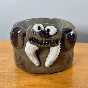 May include: A brown ceramic cup featuring a sculpted walrus face on the front. The walrus has white tusks, white eyes with black pupils, a black nose, and dark brown whiskers. Two dark brown flippers are attached to the sides of the cup.