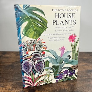 以下が含まれることがあります： ラッセル・C・モット著、アラン・シンガー挿絵のハードカバー本「The Total Book of House Plants」。表紙には、大きな緑のモンステラの葉、ピンクのサボテンの花、そして様々な観葉植物がフルカラーで描かれています。