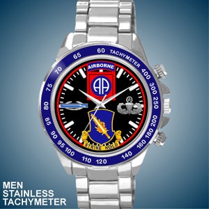以下が含まれることがあります： 黒い文字盤に「AIRBORNE」と「STRIKE HOLD」の文字と軍事記章が描かれた、シルバーのステンレススチール製腕時計。タキメーターの目盛りが付いたブルーのベゼルとシルバーのバンドが付いています。「MEN STAINLESS TACHYMETER」の文字が下部にあります。