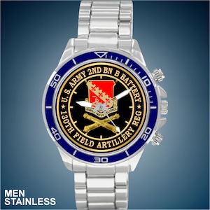 May include: A silver watch with a blue bezel and a white face. The face features a gold crest with the text "U.S. ARMY 2ND BN B BATTERY ★ 130TH FIELD ARTILLERY REG" and the words "SEMPLA PARVO ELIORES".