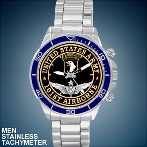 May include: A silver watch with a black face and blue bezel. The face features a gold eagle with wings spread, a star, and the text "United States Army 101st Airborne".