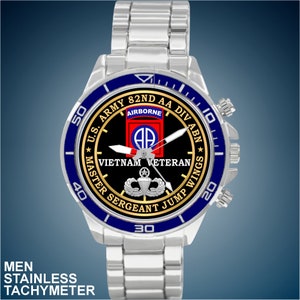 May include: A silver stainless steel watch with a black face and blue bezel. The face features the text "U.S. ARMY 82ND AA DIV ABN", "AIRBORNE", "VIETNAM VETERAN", "MASTER SERGEANT JUMP WINGS", and a military insignia.