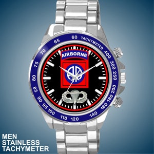 Op de afbeelding: Een zilveren roestvrijstalen horloge met een zwarte wijzerplaat met het woord "AIRBORNE" en een militair embleem. Het horloge heeft een blauwe lunette met tachymeter markeringen en een zilveren band. De tekst "MEN STAINLESS TACHYMETER" staat onderaan.