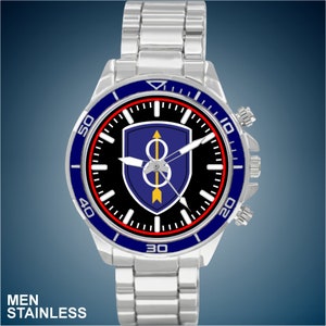 May include: A silver watch with a black face and a blue and red bezel. The watch face features a military unit insignia with a blue shield, a gold arrow, and a gold circle with a number 8 inside. The watch has a stainless steel band.