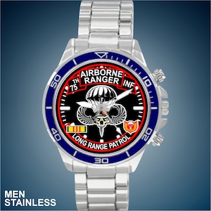 May include: A silver watch with a black face and blue bezel. The face features a skull and crossbones with wings, a parachute, and the text "Airborne Ranger Inf 75th Long Range Patrol".
