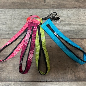 May include: Four fabric wristlet keychains with zipped pockets, featuring patterns in bright pink, dark magenta, lime green, and turquoise blue. Each keychain includes a black zip, a black fabric lining, and a silver-toned metal swivel clasp.