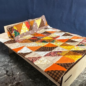 May include: A miniature wooden bed frame featuring a patchwork quilt and two matching pillows. The bedding displays a geometric pattern of triangles in shades of orange, yellow, brown, and cream, with various textures and small printed motifs.