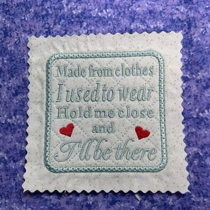 May include: A white fabric square with a blue embroidered border and text that reads "Made from clothes I used to wear Hold me close and I'll be there". Two red embroidered hearts are on either side of the word "and".