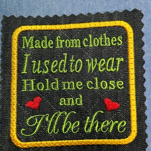 May include: Black fabric patch with a yellow border and green embroidered text that reads "Made from clothes I used to wear Hold me close and I'll be there". Two red hearts are embroidered on either side of the word "and".