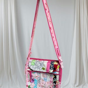 May include: A pink patchwork crossbody bag with a floral print strap. The bag is made of various fabrics and colours, including pink, blue, green, and white.