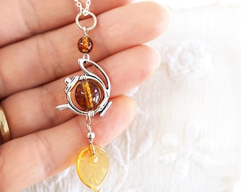 May include: A silver teapot charm necklace with amber glass beads. The charm features a detailed teapot design with a small amber glass bead in the center. The necklace is on a silver chain and has a small amber glass leaf pendant.