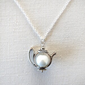 May include: A silver teapot necklace with a white pearl inside. The chain is silver and the teapot is detailed with a spout and handle.