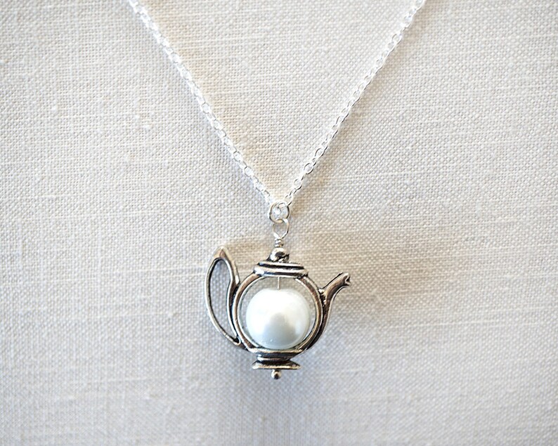 May include: A silver teapot necklace with a white pearl inside. The chain is silver and the teapot is detailed with a spout and handle.